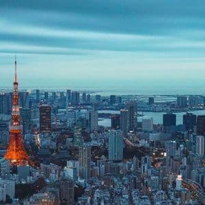 La Torre di Tokyo illuminata di arancione si staglia contro il denso paesaggio urbano al tramonto, con una baia visibile sullo sfondo.