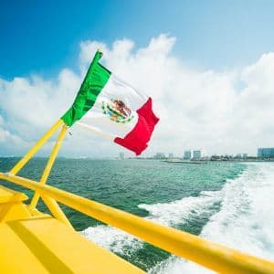 A Mexican flag waving from the back of a yellow boat on the sea, with a wake trailing behind and a city skyline in the distance.