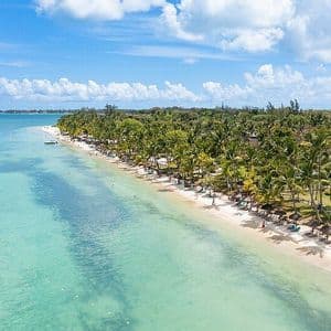 Vista aerea di una spiaggia tropicale con acqua turchese, sabbia bianca e palme sotto un cielo azzurro e nuvoloso.