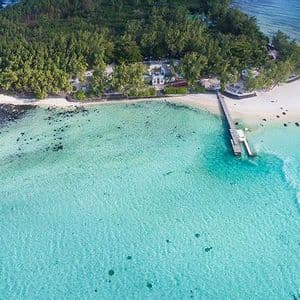 Una vista aerea di una piccola isola ricoperta di alberi, con una spiaggia sabbiosa e un molo, circondata da acqua cristallina turchese con un motoscafo che la attraversa.