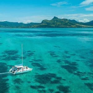 Vista aérea de un catamarán con gente a bordo navegando en aguas turquesas y cristalinas, con una isla montañosa verde a lo lejos.