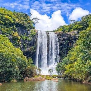 Una amplia cascada se precipita por un acantilado rocoso hasta una poza de agua, rodeada de un frondoso bosque verde bajo un cielo azul con nubes.