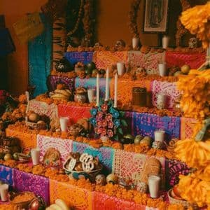 A multi-tiered Day of the Dead altar decorated with orange marigolds, skulls, candles, food, and colorful papel picado.