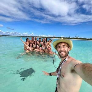 A man in a hat takes a selfie with a WeRoad group trip and a stingray in shallow turquoise water.
