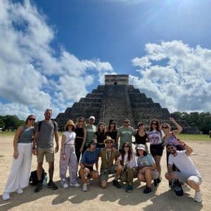 Un viaggio di gruppo WeRoad in posa per una foto davanti a una grande piramide di pietra sotto un cielo soleggiato con nuvole sparse.