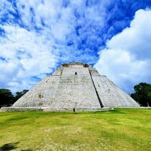 Vista dal basso di una grande piramide di pietra con una persona alla base, su un campo erboso e un cielo blu con nuvole.