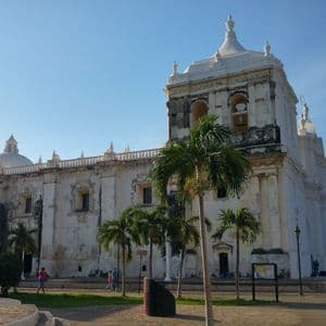 Eine große, verwitterte weiße Kathedrale mit einem Glockenturm steht auf einem Platz mit Palmen unter einem klaren blauen Himmel.