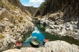 Drei kleine Ruderboote liegen am felsigen Ufer eines ruhigen Flusses, der sich durch eine sonnendurchflutete Schlucht windet, wobei sich Wolken im Wasser spiegeln.