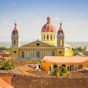 Eine große gelbe Kathedrale mit roten Kuppeln thront über einer Stadtlandschaft aus Terrakottadächern, mit einem großen Gewässer im Hintergrund.
