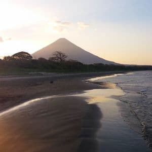 Ein weißer Reiher watet in den flachen Wellen eines dunklen Sandstrandes, wobei sich ein großer Vulkan vor dem Sonnenuntergangshimmel abzeichnet.
