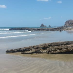 Ein Sandstrand mit großen, dunklen Felsformationen am Ufer und im Meer unter einem klaren blauen Himmel.