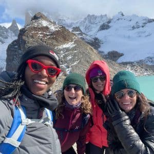 A WeRoad group trip of four women smiling for a selfie in front of a turquoise lake and snow-capped mountains.