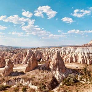 A wide landscape of conical rock formations in a dry valley under a bright blue sky with clouds.