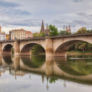 Un puente de piedra con múltiples arcos cruza un río tranquilo, reflejando el paisaje urbano histórico y el cielo nublado en su superficie.