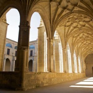 La lumière du soleil traverse les arches gothiques d'un cloître en pierre, projetant de longues ombres dans le couloir au plafond voûté orné.