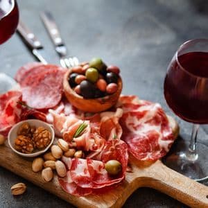 Une planche de charcuterie avec des viandes salées, des olives, des noix et deux verres de vin rouge sur une surface sombre.