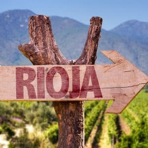Un panneau fléché en bois avec 'RIOJA' écrit en rouge pointe vers la droite, devant un vignoble verdoyant et des montagnes lointaines.