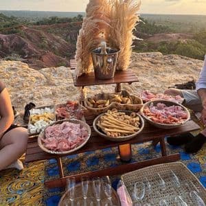 An outdoor picnic spread with sparkling wine, charcuterie, and cheese on a wooden table overlooking a canyon at sunset.