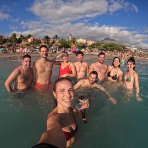 Una donna si scatta un selfie con il suo gruppo WeRoad mentre nuota nel mare su una spiaggia affollata sotto un cielo parzialmente nuvoloso.