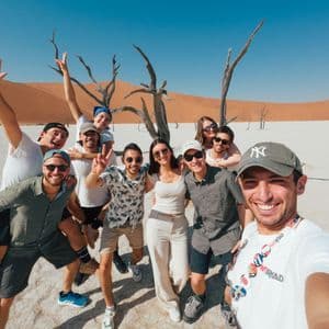Un gruppo WeRoad si fa un selfie in un paesaggio desertico con alberi secchi e grandi dune di sabbia arancioni sullo sfondo.