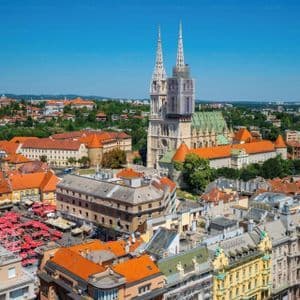 An aerial view of a historic city with red-tiled roofs, centered on a large cathedral with two spires under a clear blue sky.