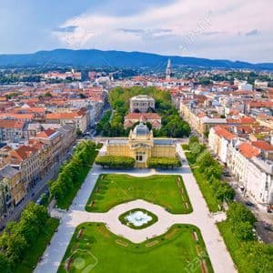 An aerial view of a large European city centered on a formal park with a fountain, leading to a grand yellow building with a dome.