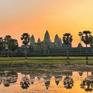 An ancient temple with tall spires and palm trees reflected in a calm body of water against an orange sky at sunrise.