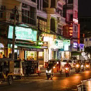A busy city street at night filled with motorcycles, tuk-tuks, and illuminated by colorful neon signs on the buildings.