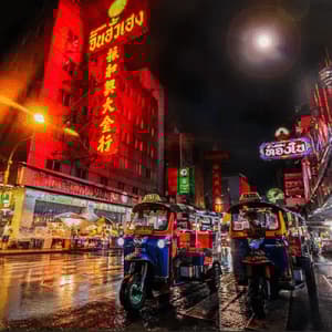Two tuk-tuks are parked on a wet city street at night, illuminated by bright red and colorful neon signs on buildings.