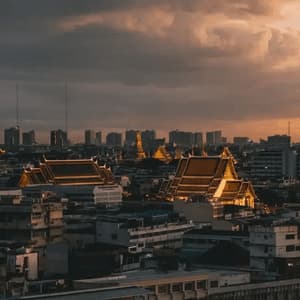 An urban skyline with ornate temple roofs in the foreground, illuminated by a warm sunset under a cloudy sky.