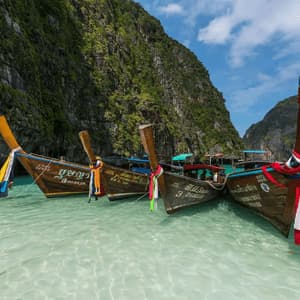 Traditional long-tail boats with colorful ribbons float on clear turquoise water beside a large, green-covered cliff.