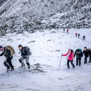 Un viaje en grupo de WeRoad haciendo senderismo en fila india por una ladera de montaña nevada, usando bastones de trekking y llevando mochilas.