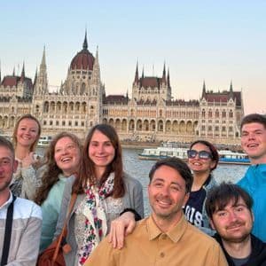 Un voyage de groupe WeRoad souriant ensemble pour une photo devant un grand bâtiment gouvernemental orné sur une rivière au crépuscule.