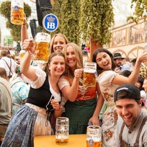 A WeRoad group trip of friends in traditional Bavarian outfits smiling and raising their beer mugs in a crowded beer hall.