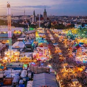 An aerial view of a crowded amusement park at dusk, with colorful lights illuminating rides and stalls against a city skyline.