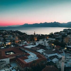 Aerial view of a coastal city with minarets overlooking a bay at sunset, with mountains in the distance.