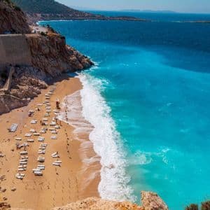 An aerial view of a sandy beach with white lounge chairs next to turquoise ocean waves at the bottom of a rocky cliff.