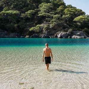 A man in black shorts, seen from behind, wades into shallow, clear turquoise water towards a tree-covered shoreline.