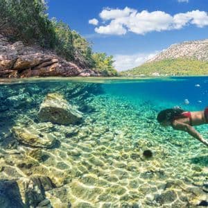 A split-level view of a woman in a red bikini swimming underwater in clear turquoise sea, with a rocky, tree-lined coast visible above the surface.