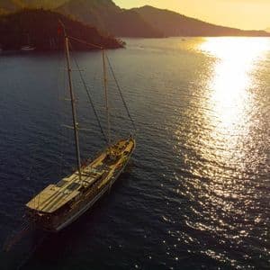 An aerial view of a wooden gulet sailboat anchored in a calm bay at sunset, with golden light reflecting on the water.