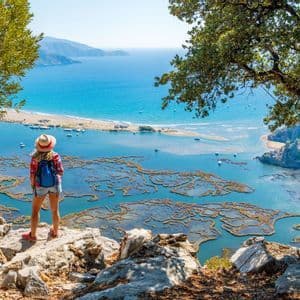 A person with a backpack and hat stands on a rocky clifftop, looking out over a coastal delta and a wide blue sea.