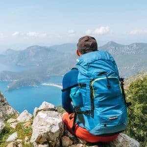 A hiker with a blue backpack sits on a rocky cliff, looking out over a coastal landscape of islands and mountains.