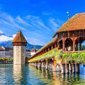 A wooden covered bridge and stone tower stand on a river, with city buildings and distant mountains under a bright blue sky.
