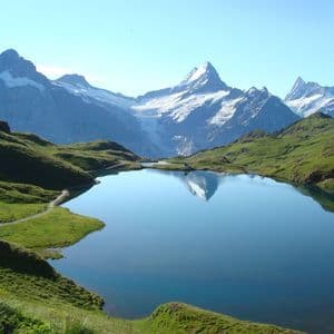 A still alpine lake reflects a snow-capped mountain peak, surrounded by green hills under a clear blue sky.