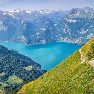A hiker with a red backpack descends a steep mountain path overlooking a turquoise lake and distant snow-capped mountains.