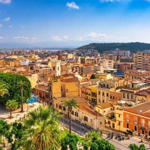 Una vista aérea de una ciudad costera soleada con edificios de colores cálidos, palmeras y colinas distantes bajo un cielo azul.