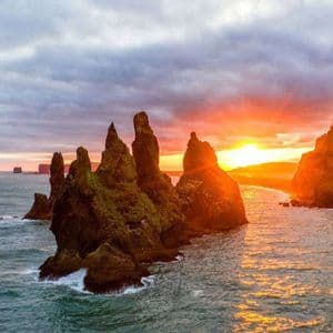 Zackige, moosbewachsene Felsnadeln ragen aus dem Meer, während die Sonne untergeht und ein orangefarbenes Glühen auf das Wasser wirft.