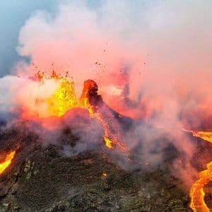 Un volcan actif entre en éruption, avec de la lave en fusion s'écoulant en rivières le long de son flanc sombre et rocheux sous un ciel rempli de fumée.