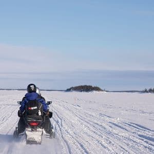 Rückansicht von zwei Personen einer WeRoad Gruppenreise, die mit einem Schneemobil durch eine weite, verschneite Landschaft unter einem klaren blauen Himmel fahren.