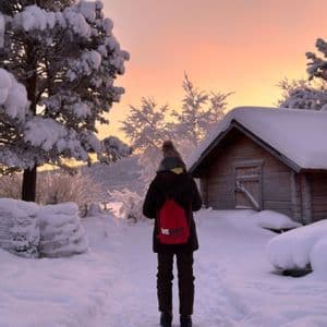 Una persona de espaldas, llevando una mochila WeRoad roja, se encuentra en un sendero nevado junto a una cabaña de madera al atardecer.